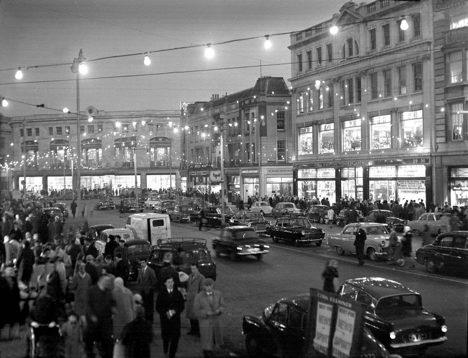 Shoppers hurry along the pavements on either side of a busy street. Bare bulb Christmas decorations hang overhead. Cars parked on both side of the road.